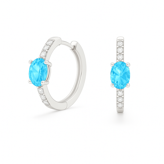 Elegant oval-cut Swiss blue topaz huggie earrings in white gold-plated 925 silver with sparkling Moissanite accents for a fresh charm. Available at BSI Jewelry.