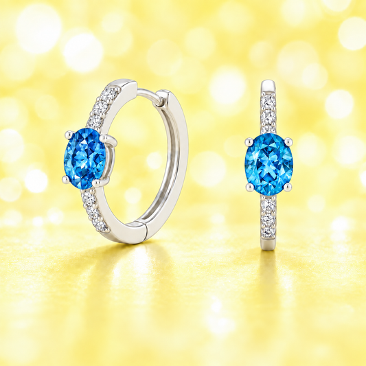 Elegant oval-cut Swiss blue topaz huggie earrings in white gold-plated 925 silver with sparkling Moissanite accents for a fresh charm. Available at BSI Jewelry.